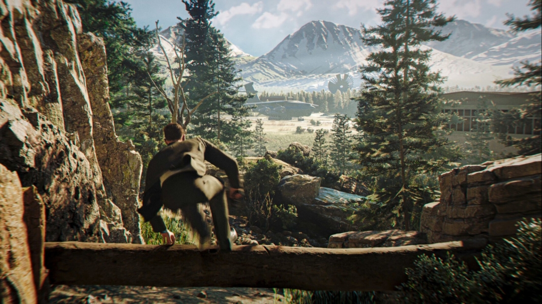 Person in suit jumping over a log in a forested mountain landscape with snow-capped peaks and buildings in the valley