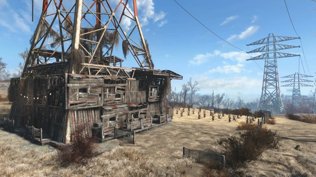 Dilapidated wooden and metal building beneath a power transmission tower in a sparse landscape with scattered vegetation and blue sky.