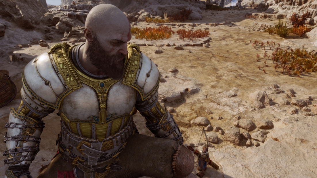 A muscular man in white and gold armor stands on rocky, dry ground with sparse vegetation