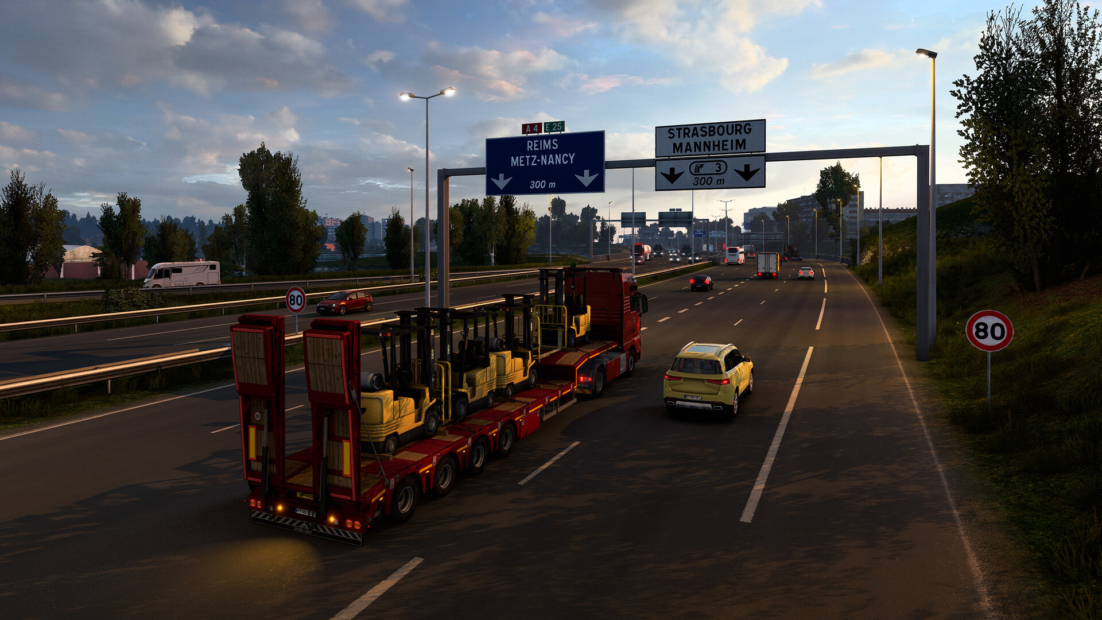 Red and black tanker truck driving on a multi-lane city road in Euro Truck Simulator 2, surrounded by cars and buildings under a partly cloudy sky