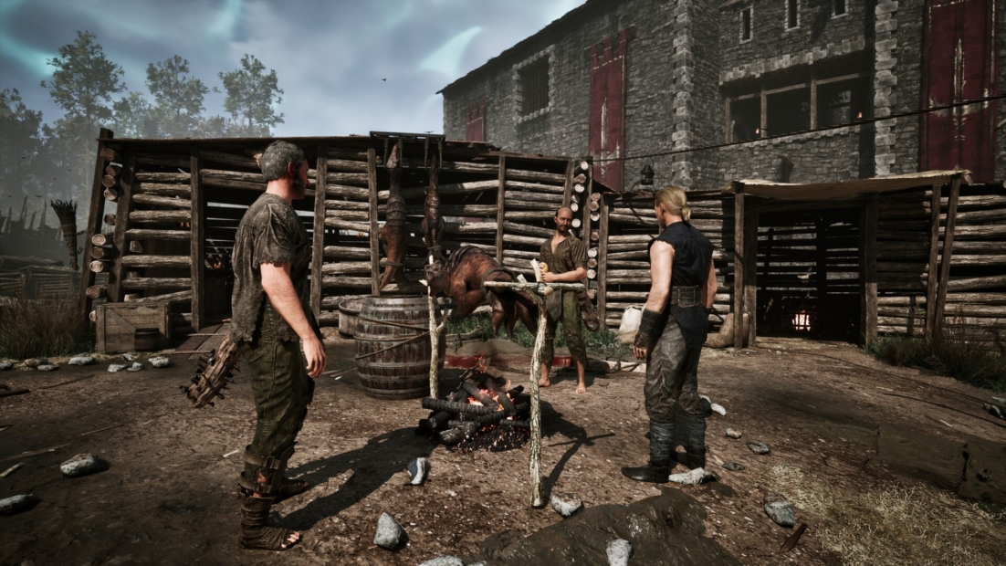 Three men in medieval clothing stand around a campfire in front of a wooden barricade and a stone building