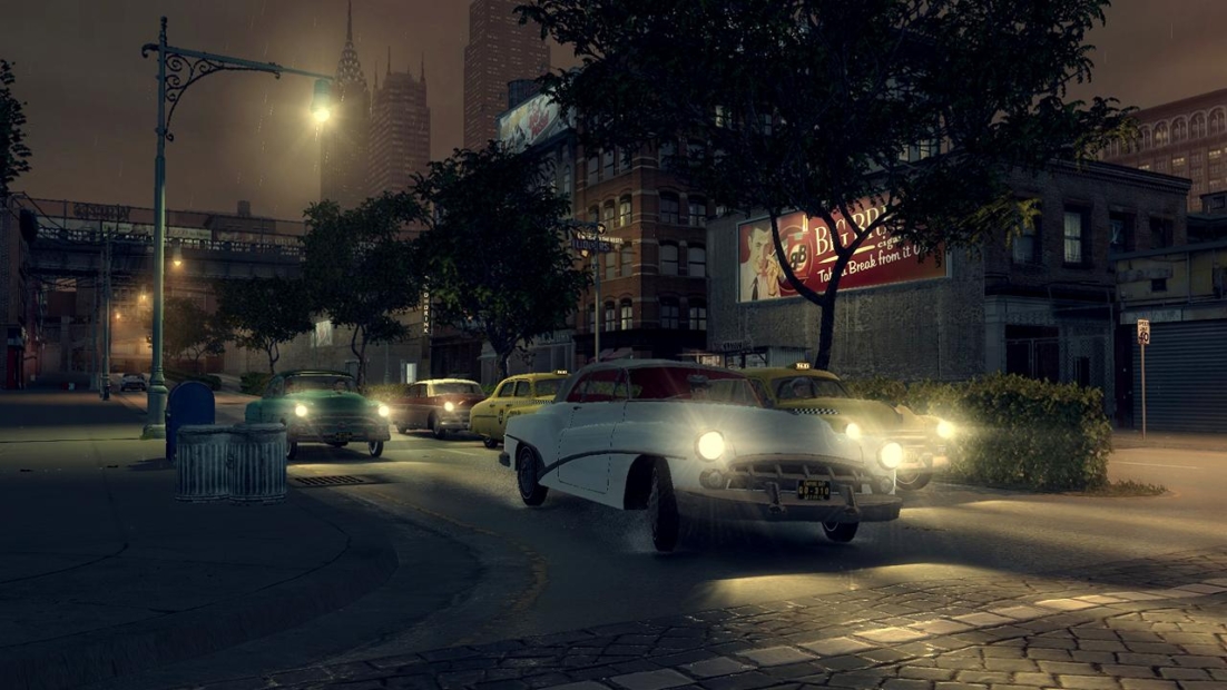 Night street scene from Mafia II (2011) showing vintage cars driving on a city road with buildings and streetlights in the background