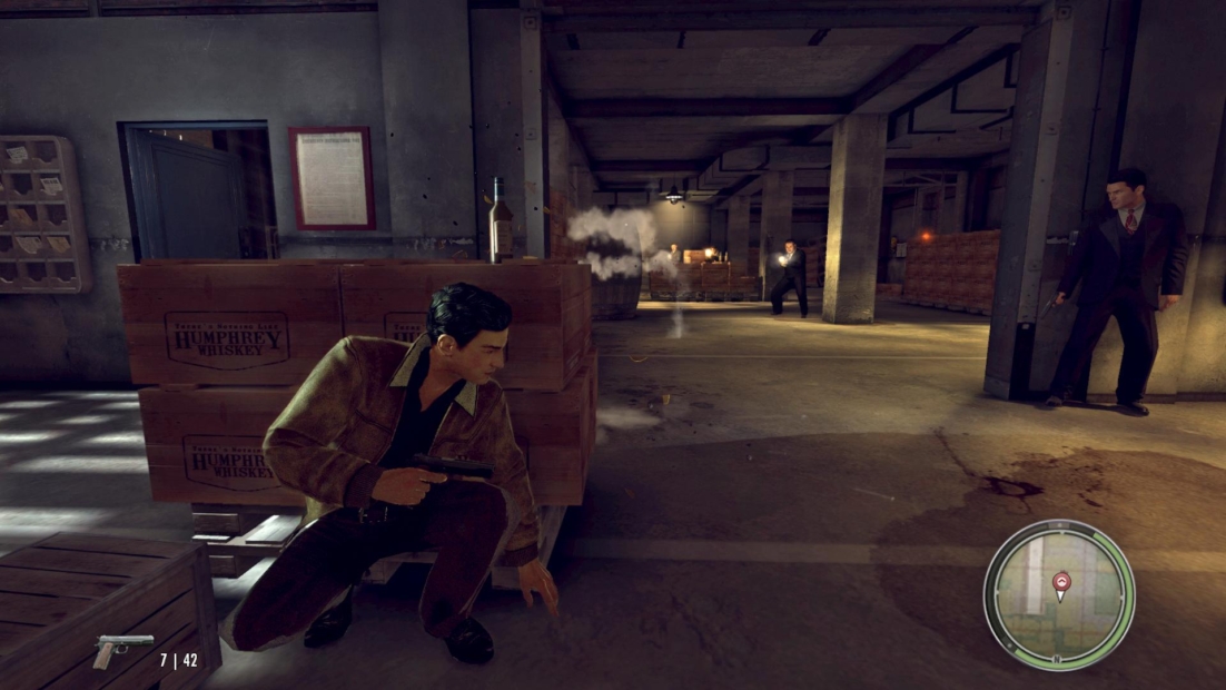 Character crouching behind wooden crates labeled 'Humphrey Whiskey' holding a pistol in a dimly lit warehouse with two armed men in the background, from the game Mafia II