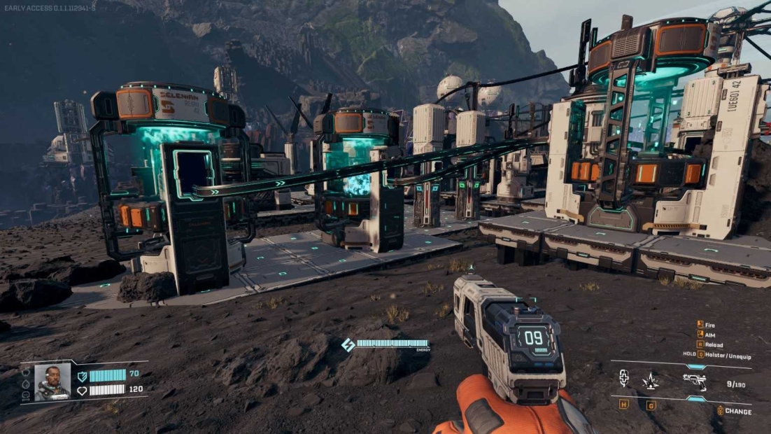 Several futuristic machines with glowing blue elements stand on a rocky surface connected by pipes, a hand holding a device in the foreground, mountains in the background.