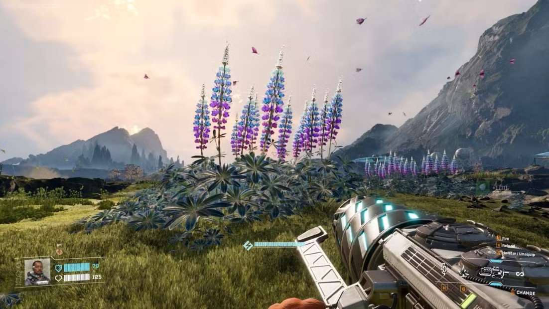 Blick auf eine Wiese mit hohen, spitzen Blütenständen vor Berglandschaft, im Vordergrund eine futuristische Waffe in einer Ego-Perspektive